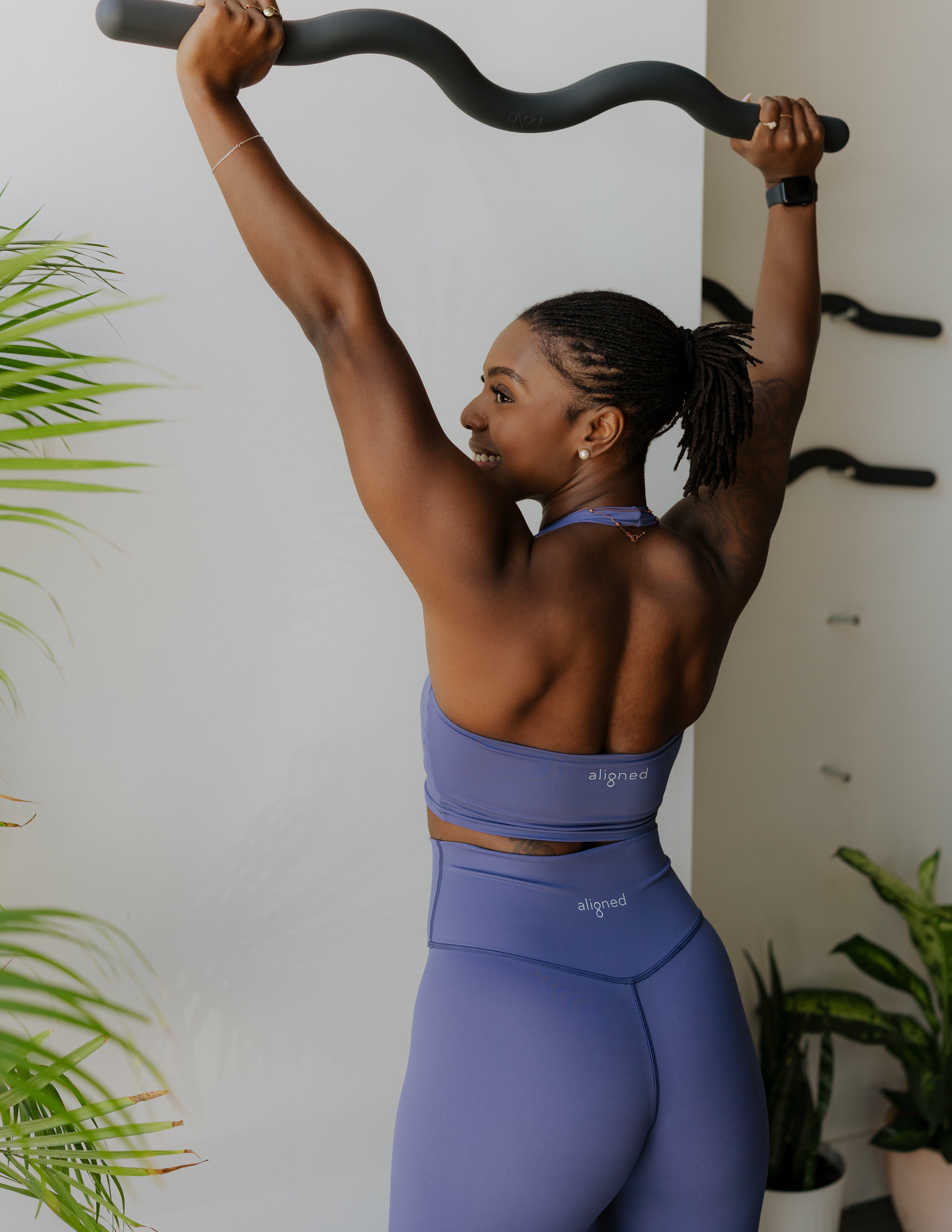 Person in purple athletic wear exercising with a barbell against a white wall with plants.
