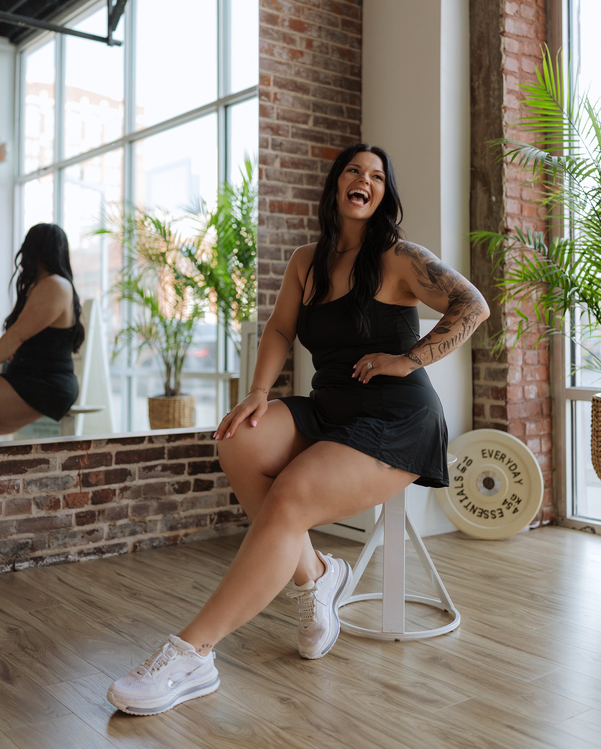 Woman in a black dress sitting on a white stool in a modern interior setting with large windows and plants.